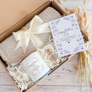 May include: A gift box filled with a beige throw blanket, a white candle with a floral design and the text "Happy Mother's Day", a small glass jar with matches, and a card with the text "Happy Mother's Day" and a floral design.