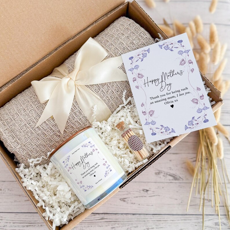 May include: A gift box filled with a beige throw blanket, a white candle with a floral design and the text "Happy Mother's Day", a small glass jar with matches, and a card with the text "Happy Mother's Day" and a floral design.