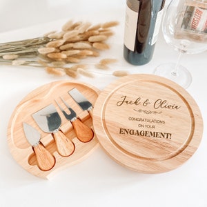 May include: A wooden cheese board with a round top and a separate compartment for cheese knives. The board is engraved with the text "Jack & Olivia Congratulations on your ENGAGEMENT!"