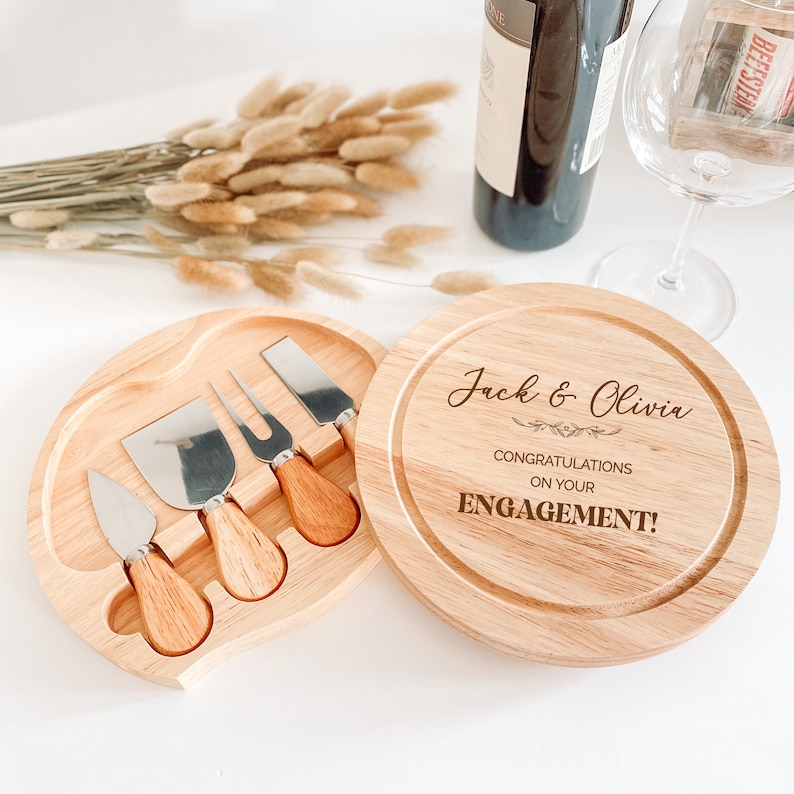 May include: A wooden cheese board with a round top and a separate compartment for cheese knives. The board is engraved with the text "Jack & Olivia Congratulations on your ENGAGEMENT!"