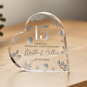 Könnte beinhalten: Ein transparentes, herzförmiges Kristall-Andenken mit silbernem Blumendesign. Die Zahl 15 ist in Silber gedruckt, zusammen mit dem Text "Kristall-Hochzeitsjubiläum Martha & Arthur 23.05.2010".