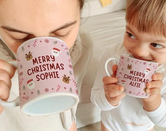 Könnte beinhalten: Zwei rosa Weihnachtstassen mit weißen Henkeln. Auf einer Tasse steht "MERRY CHRISTMAS SOPHIE" und auf der anderen "MERRY CHRISTMAS ALFIE". Die Tassen sind mit Weihnachtsmann, Rentieren, Schneeflocken und Zuckerstangen verziert.