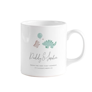 May include: White ceramic mug featuring a whimsical design with two cartoon dinosaurs, a balloon, and the text "Daddy & Sophie." The mug also includes the phrase "FROM THE VERY FIRST MOMENT, IT'S ALWAYS BEEN US."