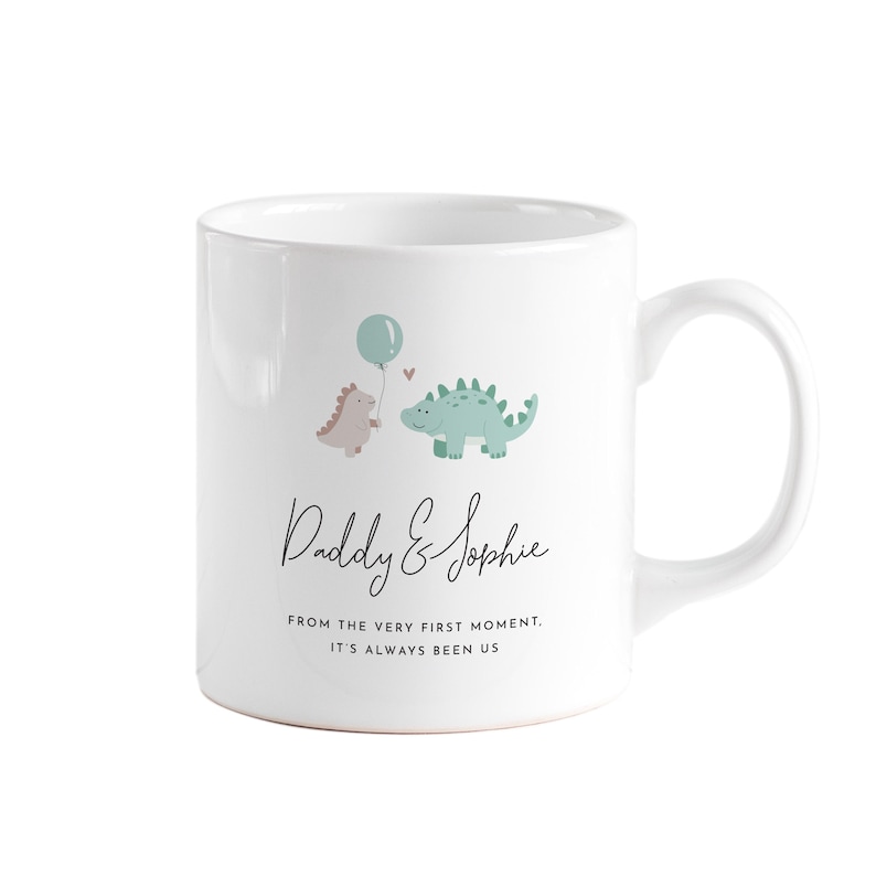 May include: White ceramic mug featuring a whimsical design with two cartoon dinosaurs, a balloon, and the text "Daddy & Sophie." The mug also includes the phrase "FROM THE VERY FIRST MOMENT, IT'S ALWAYS BEEN US."