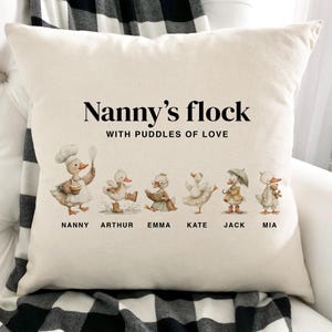 May include: A cream-coloured cushion featuring the words "Nanny's flock WITH PUDDLES OF LOVE." Below the text are six cartoon ducks, each with a different outfit and name. The names are Nanny, Arthur, Emma, Kate, Jack, and Mia.
