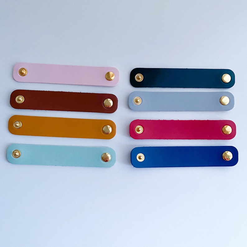 K&ouml;nnte beinhalten: Eine Sammlung von acht Leder-Kabelorganisatoren in verschiedenen Farben, darunter Rosa, Braun, Gelb und Blau. Jeder rechteckige Organizer ist mit zwei goldfarbenen Nieten befestigt.