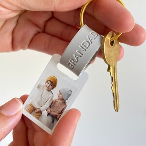 Puede incluir: Un llavero de foto blanco con una imagen de dos ni&ntilde;os con sombreros. El llavero tiene una correa de cuero gris con la palabra "GRANDAD" grabada en relieve. Una llave dorada est&aacute; unida al llavero.