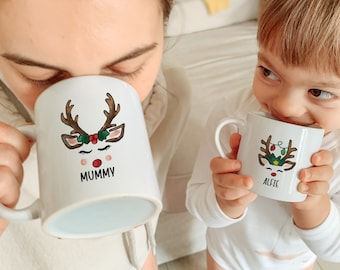 Könnte beinhalten: Zwei weiße Keramikbecher mit Rentiermotiven. Der größere Becher sagt "Mummy" und der kleinere Becher sagt "Alfie".