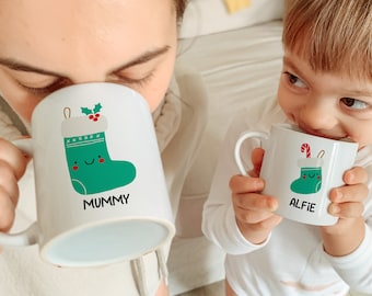 Könnte beinhalten: Zwei weiße Tassen mit Weihnachtsstrumpf-Designs. Die Tasse links sagt "Mummy" und die Tasse rechts sagt "Alfie".