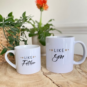 May include: Two white ceramic mugs with black text. The smaller mug says "Like Father" with gold stars. The larger mug says "Like Son" with gold stars. The mugs are on a wooden surface.
