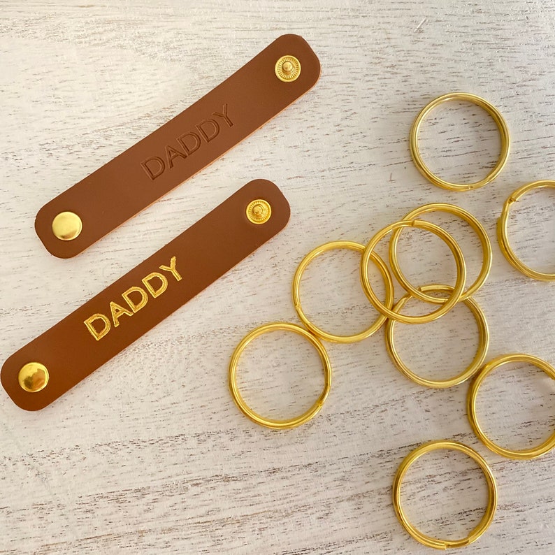 K&ouml;nnte beinhalten: Zwei braune Leder-Schl&uuml;sselanh&auml;nger mit dem Wort "DADDY" in goldfarbenen Buchstaben gepr&auml;gt. Jeder Schl&uuml;sselanh&auml;nger hat einen goldenen Druckknopfverschluss. Mehrere goldfarbene Schl&uuml;sselringe sind ebenfalls sichtbar.