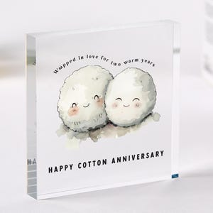 Op de afbeelding: Een witte acrylblok met een aquarel-illustratie van twee pluche witte wattenbolletjes met blije gezichten. De tekst "Wrapped in love for two warm years" staat boven de wattenbolletjes en "Happy Cotton Anniversary" staat onder de wattenbolletjes.