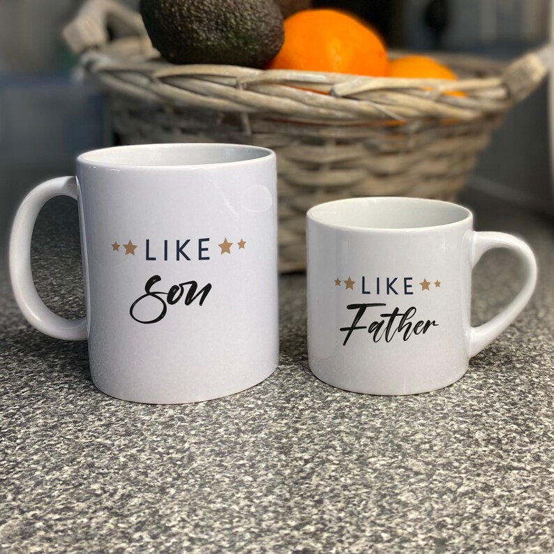 May include: Two white ceramic mugs with black text and gold stars. The larger mug says "Like Son" and the smaller mug says "Like Father".