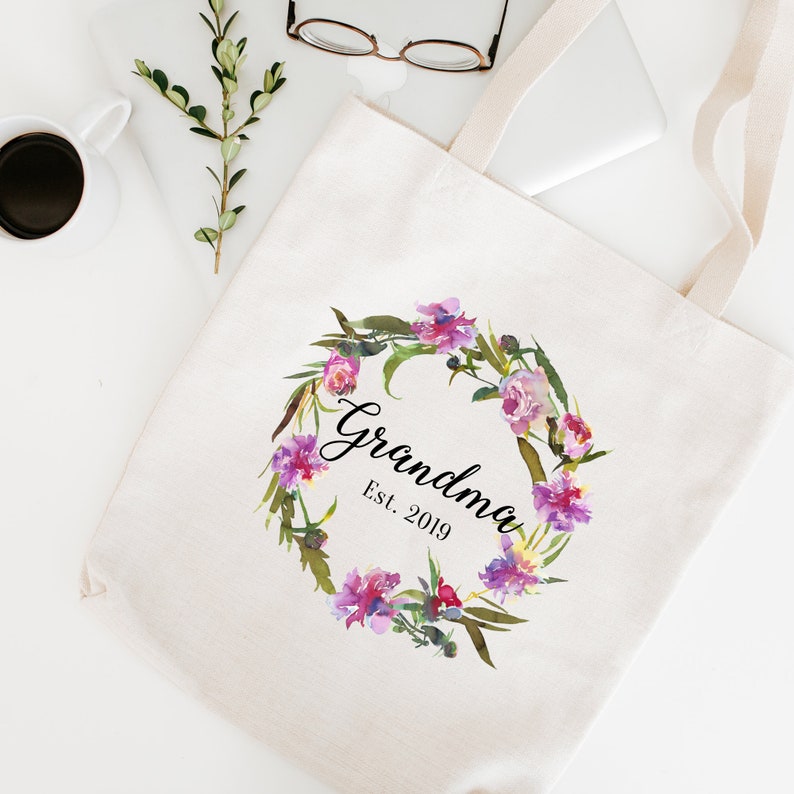 Op de afbeelding: Een witte canvas tote bag met een bloemenkransontwerp en de tekst "Grandma Est. 2019".