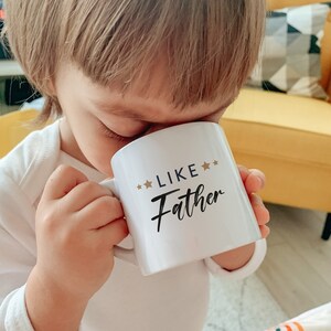 May include: White ceramic mug with black text that reads "Like Father" and gold stars. A child is drinking from the mug.