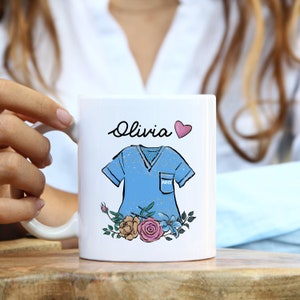 May include: White ceramic mug with a blue scrubs design, floral accents, and the text "Olivia" with a pink heart.
