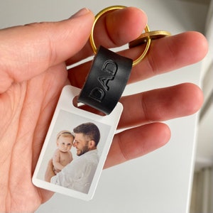 K&ouml;nnte beinhalten: Ein personalisierter Schl&uuml;sselanh&auml;nger mit einem Foto von Vater und Kind. Der Schl&uuml;sselanh&auml;nger hat einen schwarzen Lederriemen mit dem gepr&auml;gten Wort "DAD" und einen goldfarbenen Ring. Das Foto ist rechteckig und zeigt einen Mann, der ein Baby h&auml;lt.