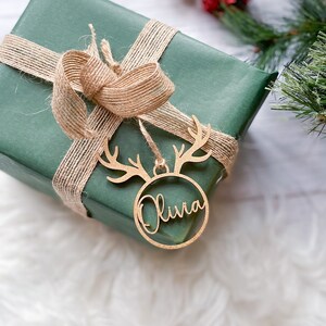 May include: A green gift box wrapped with brown twine and a wooden reindeer ornament with the name "Olivia" inscribed on it.