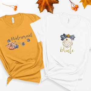 May include: Two t-shirts with a fall wedding theme. The white t-shirt says "Bride" with a gold glitter font and a white pumpkin with gold glitter dots and blue leaves. The mustard yellow t-shirt says "Bridesmaid" with a black font and a small orange pumpkin with blue leaves.