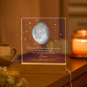 Puede incluir: Luz de noche personalizada con base de madera. El panel acrílico muestra una ilustración detallada de la luna sobre un fondo estrellado. El texto dice: "Así es como se veía la Luna el día que te convertiste en mi padre. Te amo hasta la Luna y de vuelta. Grace 12.07.1971."