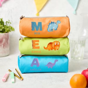 Può includere: Tre astucci impilati, arancione, verde e blu. Ogni astuccio presenta un'illustrazione di dinosauro e un nome: "MIA", "EMMA" e "AMELIA". Gli astucci sono cilindrici con chiusura a zip. Lo sfondo include matite, gomme e frutta.