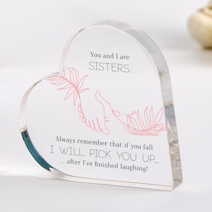 Può includere: Un blocco acrilico trasparente a forma di cuore con il testo "You and I are SISTERS." e un'illustrazione rosa di mani. Il testo "Always remember that if you fall I WILL PICK YOU UP... after I've finished laughing!" è stampato.