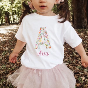 Peut inclure: T-shirt blanc avec une lettre A florale et le nom Ava imprimé en rose en dessous. Le t-shirt est porté par un enfant.