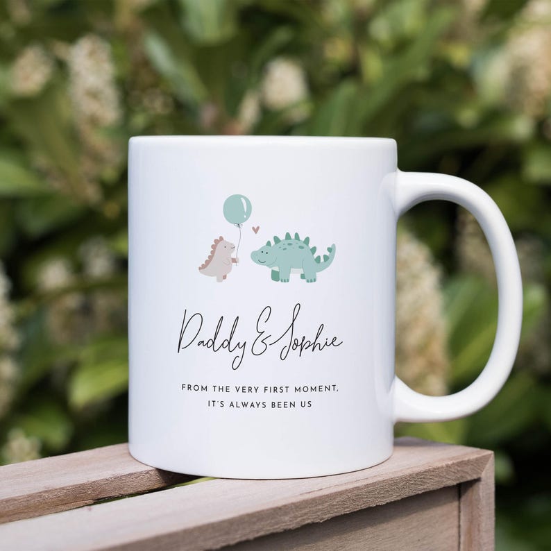 May include: White ceramic mug featuring a whimsical design with two cartoon dinosaurs, one holding a balloon. The mug has the text "Daddy & Sophie" and a sentimental quote. A perfect gift for a parent and child.