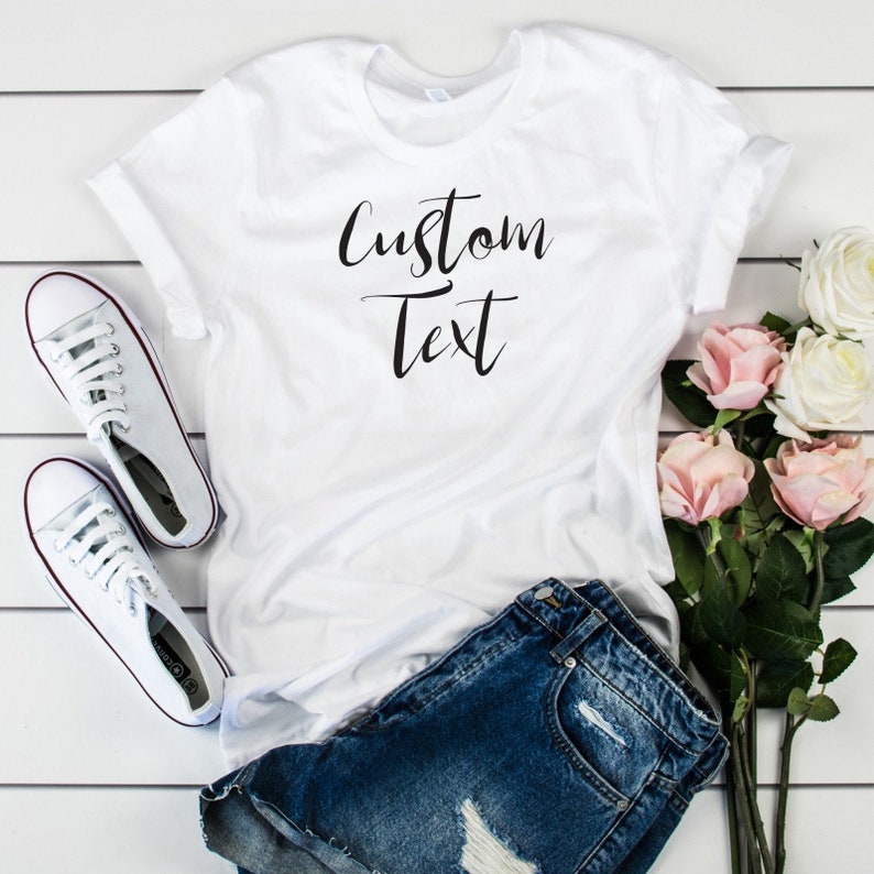 May include: White t-shirt with the text "Custom Text" in black script font. The shirt is laid flat on a white wooden surface with a pair of white sneakers and blue denim shorts.