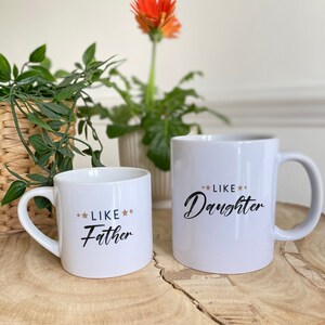 May include: Two white ceramic mugs with black text and gold stars. The smaller mug says "Like Father" and the larger mug says "Like Daughter".
