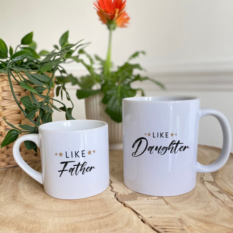 May include: Two white ceramic mugs with black text and gold stars. The smaller mug says "Like Father" and the larger mug says "Like Daughter".