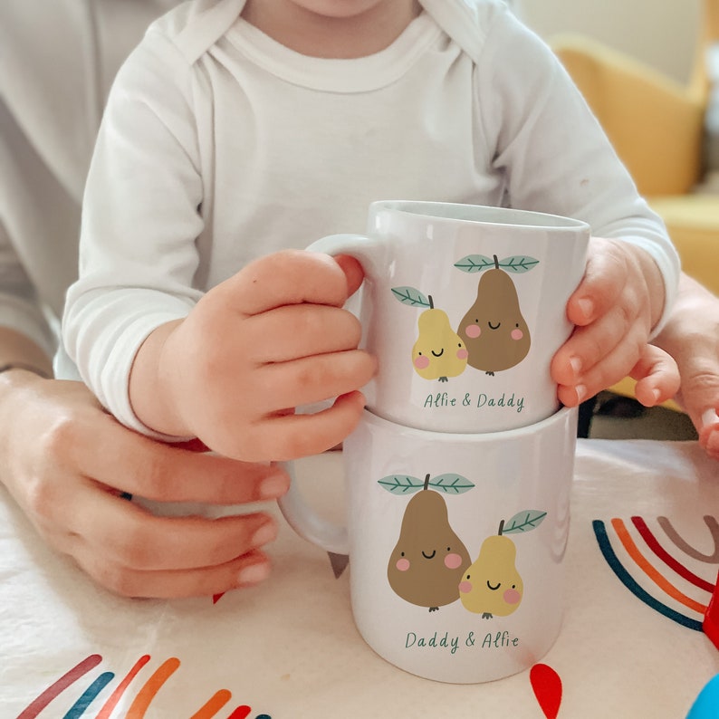 May include: Two white ceramic mugs with pear illustrations and the text "Allie & Daddy" and "Daddy & Allie" on them. The mugs are stacked on top of each other.