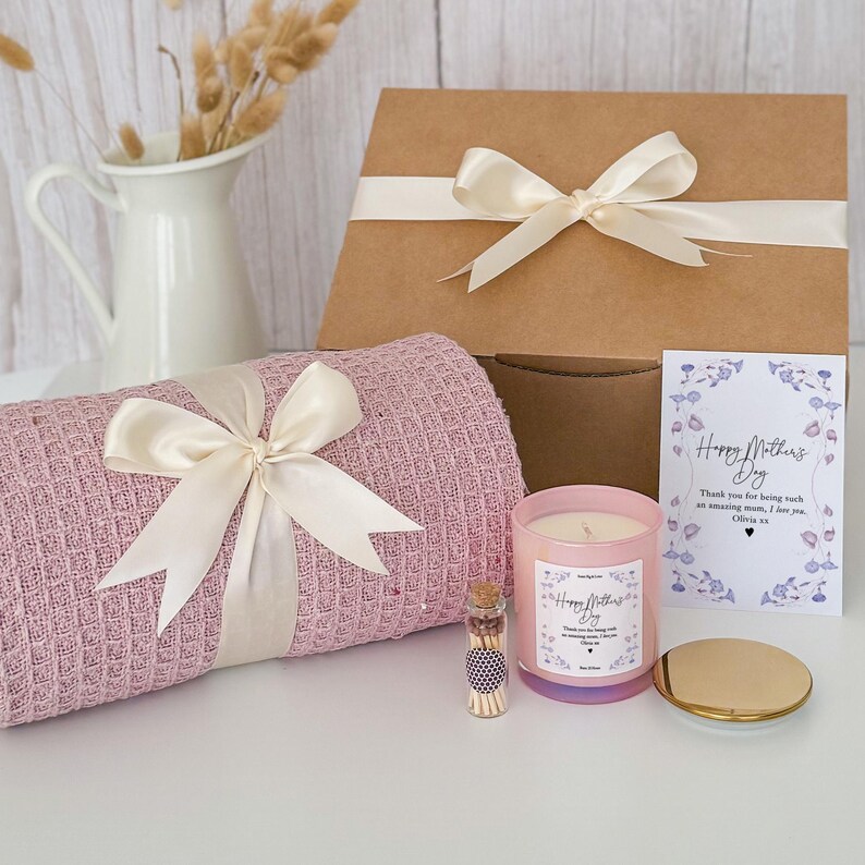 May include: A gift box set for Mother's Day with a pink throw blanket, a pink candle with a "Happy Mother's Day" label, a small glass jar of matches, and a card with the text "Happy Mother's Day. Thank you for being such an amazing mom, I love you. Olivia xx."