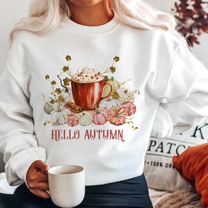 Peut inclure: Sweat-shirt blanc avec un motif graphique d'une tasse de café avec de la crème fouettée et des pépites, entourée de citrouilles et de feuilles d'automne. Le texte "HELLO AUTUMN" est imprimé sous le graphique.
