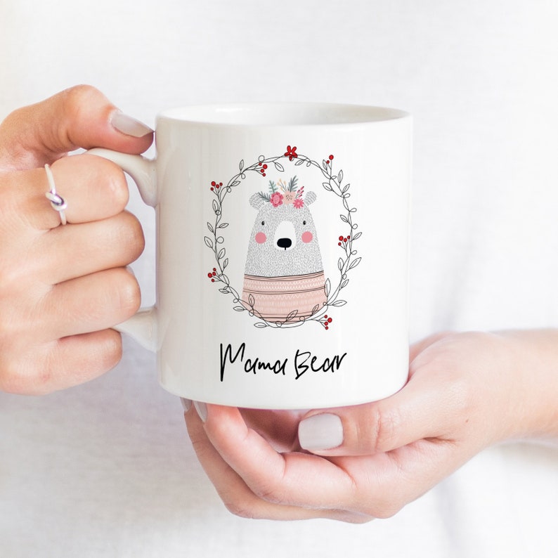 May include: White ceramic mug with a black and white illustration of a bear wearing a pink and white sweater and a floral crown. The bear is surrounded by a black wreath with red berries. The text "Mama Bear" is printed below the bear.