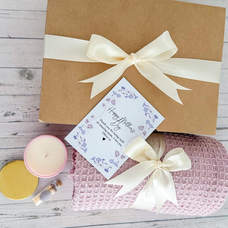 May include: A gift box with a pink blanket, a candle, and a small bottle. The box is tied with a cream ribbon and a card that says "Happy Mother's Day" with a message of thanks.