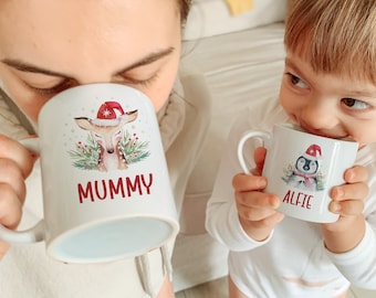 Könnte beinhalten: Zwei weiße Keramikbecher mit Weihnachtsmotiven. Der größere Becher zeigt ein Reh mit einem Weihnachtsmann-Hut und dem Text "MUMMY". Der kleinere Becher zeigt einen Pinguin mit einem Weihnachtsmann-Hut und dem Text "ALFIE".