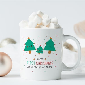 Op de afbeelding: Witte keramische mok met een groen, rood en gouden ontwerp van drie kerstboommetjes met lachende gezichtjes. De tekst "Gelukkig eerste Kerstmis als gezin met drie" staat op de mok gedrukt.