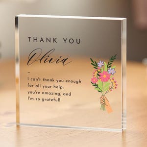 May include: A clear acrylic block with the words "THANK YOU Olivia" in black script. The block features a message of gratitude and a colourful illustration of a bouquet of flowers.