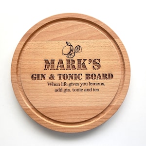 Puede incluir: Una tabla redonda de servir de madera con el texto "MARK'S GIN & TONIC BOARD" y el dicho "When life gives you lemons, add gin, tonic and ice". La tabla tiene un diseño de limón.
