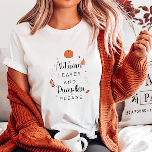 Może przedstawiać: Biała koszulka z napisem "Autumn Leaves and Pumpkin Please" czarnymi literami. Projekt zawiera małą pomarańczową dynię, liście i gwiazdki.