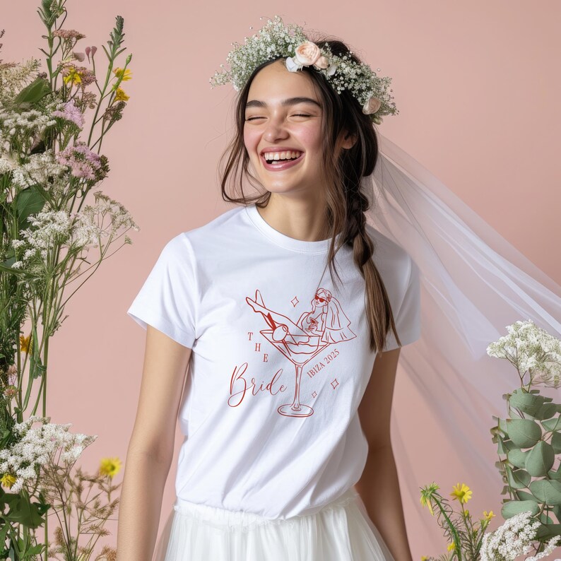 May include: A white t-shirt with a red graphic of a bride relaxing in a martini glass, with the text "The Bride Ibiza 2025". The woman is wearing a flower crown and veil. The background is a soft pink.