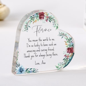 Può includere: Un ricordo in acrilico trasparente a forma di cuore con accenti floreali. Il testo recita "Florence" e un messaggio personalizzato. I bordi sono decorati con una ghirlanda di foglie verdi, bordeaux e fiori blu. Un regalo premuroso.