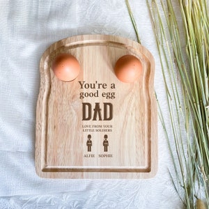 Puede incluir: Bandeja de huevos de madera con forma de rebanada de pan con dos soportes para huevos. La bandeja está grabada con el texto "You're a good egg Dad. Love from your little soldiers. Alfie Sophie."