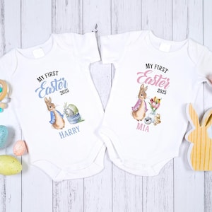 May include: Two white baby onesies with Easter designs. One has a bunny in a blue jacket, the text "My First Easter 2025" and the name "Harry". The other features a bunny in a pink scarf, the same text, and the name "Mia".