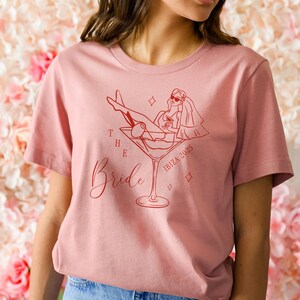 May include: A dusty rose t-shirt featuring a red graphic of a woman in a martini glass, with the words "The Bride" and "Ibiza 2025". The design includes a bride wearing sunglasses and a veil.