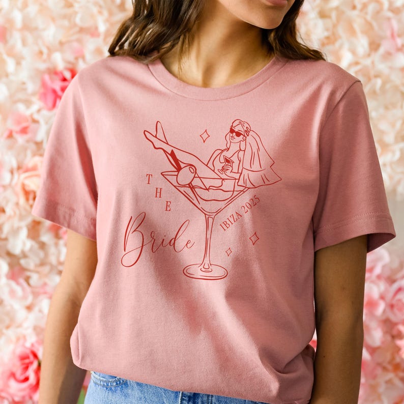 May include: A dusty rose t-shirt featuring a red graphic of a woman in a martini glass, with the words "The Bride" and "Ibiza 2025". The design includes a bride wearing sunglasses and a veil.