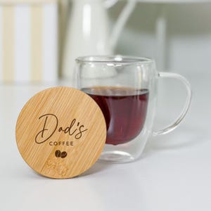 Puede incluir: Una taza de café de vidrio transparente con asa, llena de un líquido oscuro, junto a una tapa redonda de madera. La tapa está grabada con las palabras "Dadi's COFFEE" y dos iconos de granos de café.