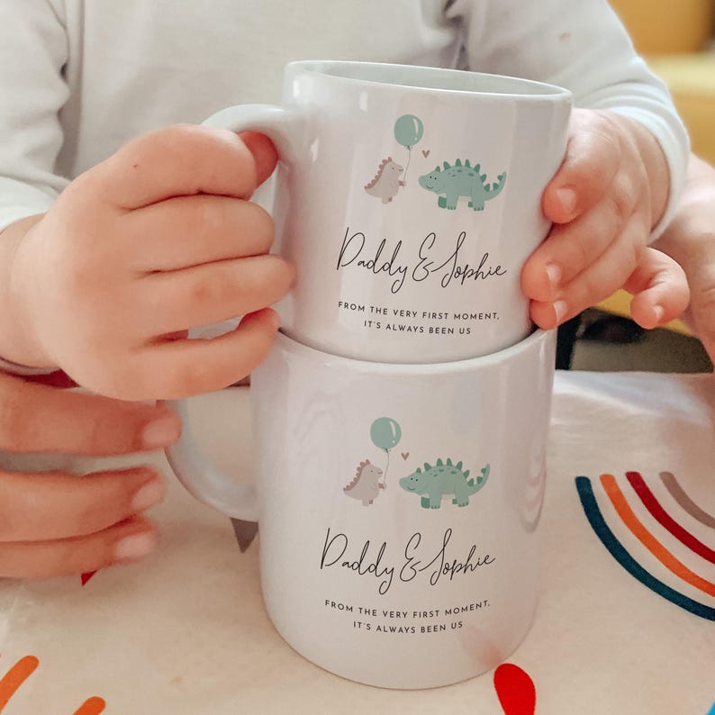 May include: Two white ceramic mugs stacked on top of each other. Each mug features a cartoon dinosaur design with a balloon and the text "Daddy & Sophie" and "FROM THE VERY FIRST MOMENT, IT'S ALWAYS BEEN US". The mugs are held by a person.