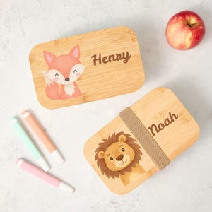 Puede incluir: Dos fiambreras de bambú con ilustraciones de animales. Una presenta un zorro y el nombre "Henry", la otra un león y el nombre "Noah". También se ven una manzana y tres rotuladores.